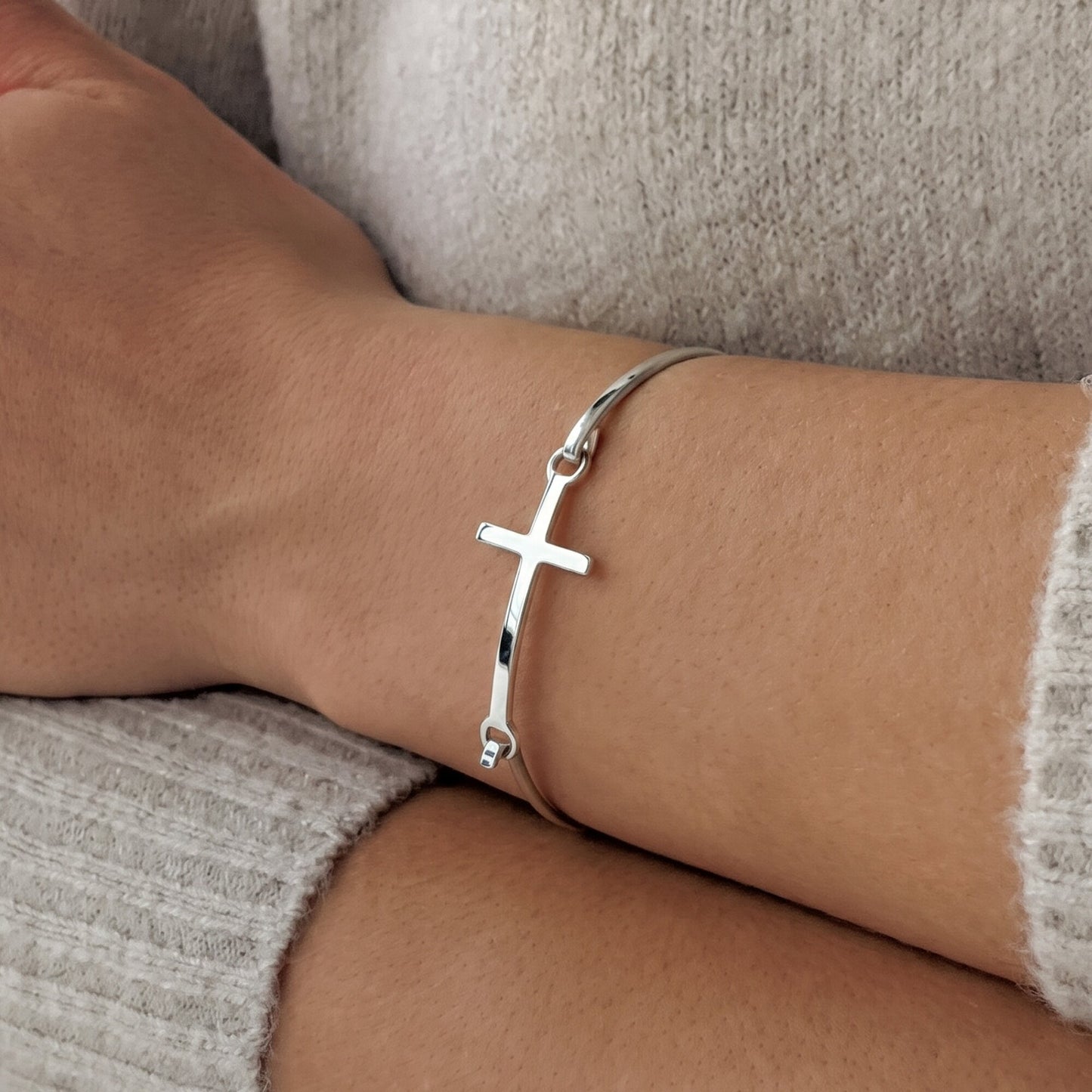 A polished sterling silver cross bangle worn on a person's wrist, shown against a soft beige knit sweater.
