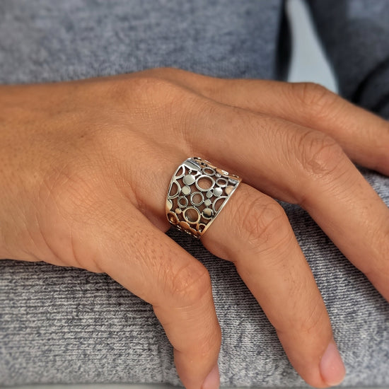 
                    
                      A close-up of a hand wearing a wide sterling silver ring with an intricate openwork circular pattern.
                    
                  