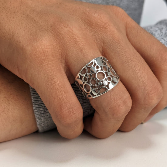 A close-up of a hand wearing a wide sterling silver ring with an openwork circular bubble pattern, resting on a white surface.