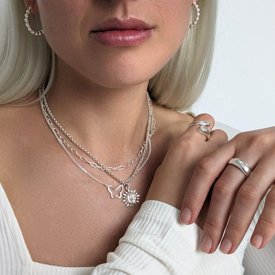 
                    
                      Sterling silver jewellery look — layered chain necklaces with butterfly and sun charms, styled with open bulbous ring, chunky band ring and beaded hoop earrings.
                    
                  